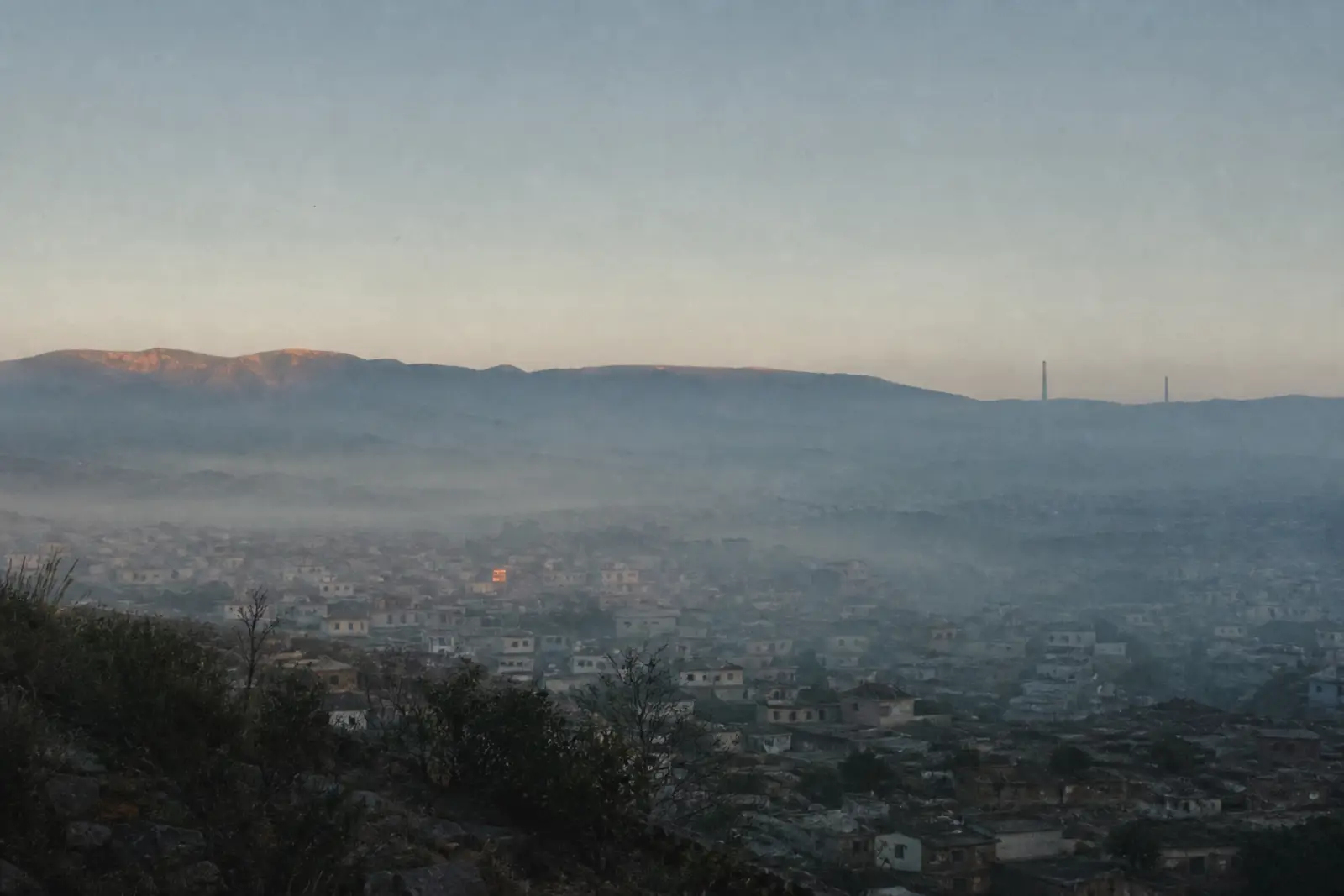 Amanhecer sobre uma cidade em um vale, com névoa baixa cobrindo os telhados e colinas ao fundo.