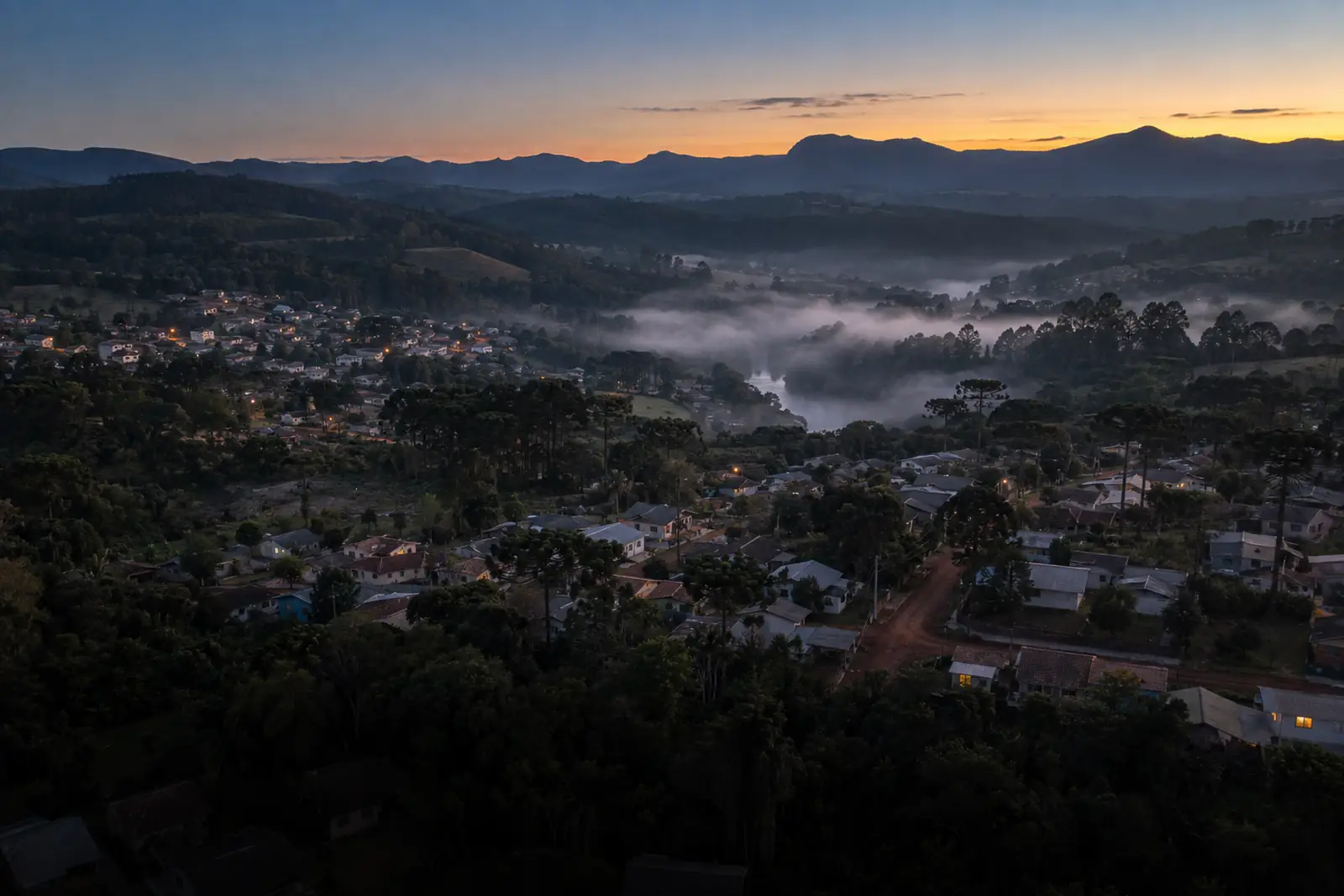 Vista aérea baixa, ao amanhecer, de uma cidade média do interior do Paraná: casas simples descendo até um vale com cerração baixa sobre um rio, araucárias entre as casas, poucas janelas acesas e uma serra verde-azulada ao fundo.