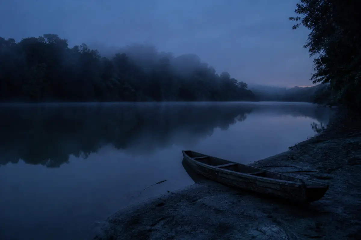 Rio largo de água barrenta ao amanhecer, com neblina sobre a superfície, mata na margem oposta e um pequeno barco de madeira encalhado na areia.