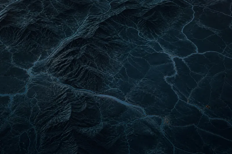 Relevo de um território em luz baixa, visto de cima: cristas e vales escuros cortados por um rio e por uma malha fina de linhas claras.