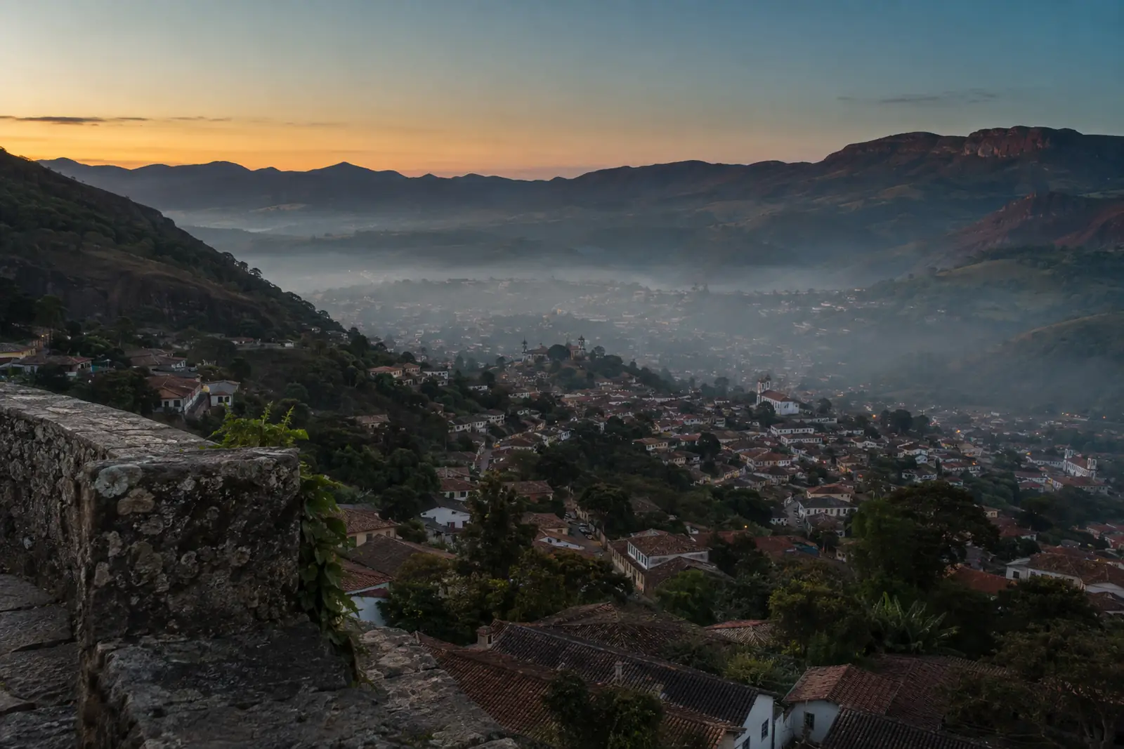 Cidade histórica de Minas Gerais vista de um mirante de pedra ao amanhecer: o casario desce pela encosta até o fundo do vale, onde uma camada fina de bruma acinzentada repousa sobre os telhados; ao fundo, serras avermelhadas e o céu clareando.