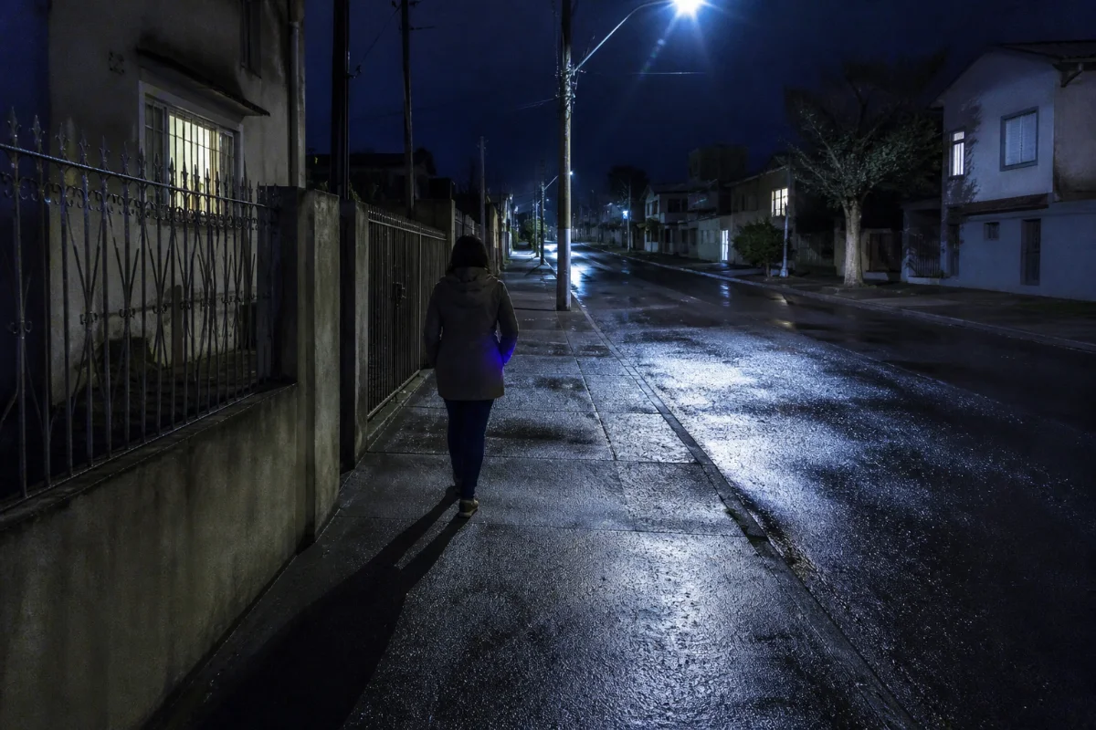 Mulher adulta caminha de costas por uma calçada de rua residencial à noite, com a mão dentro do bolso do casaco, de onde escapa a luz fraca da tela de um celular.