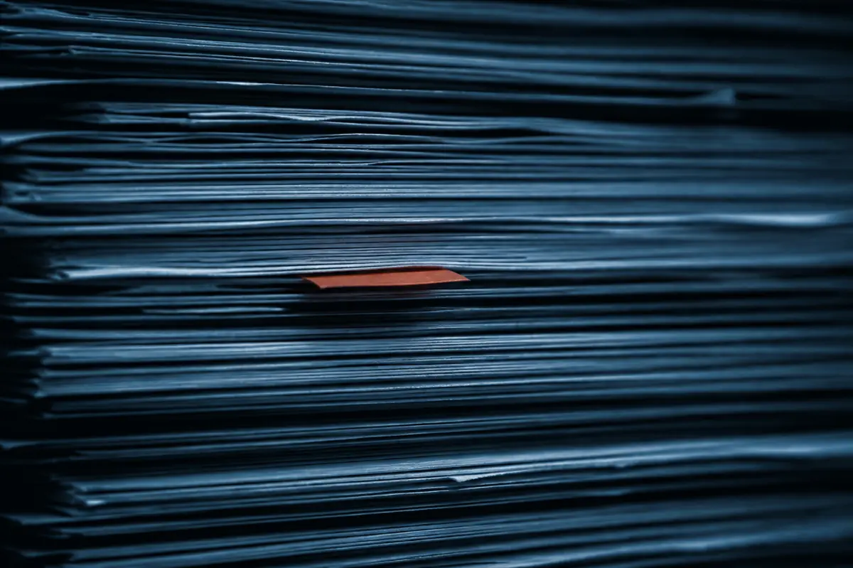 Close das bordas de uma pilha alta de documentos, formando camadas finas iluminadas de lado, com dois marcadores coloridos e um clipe metálico.